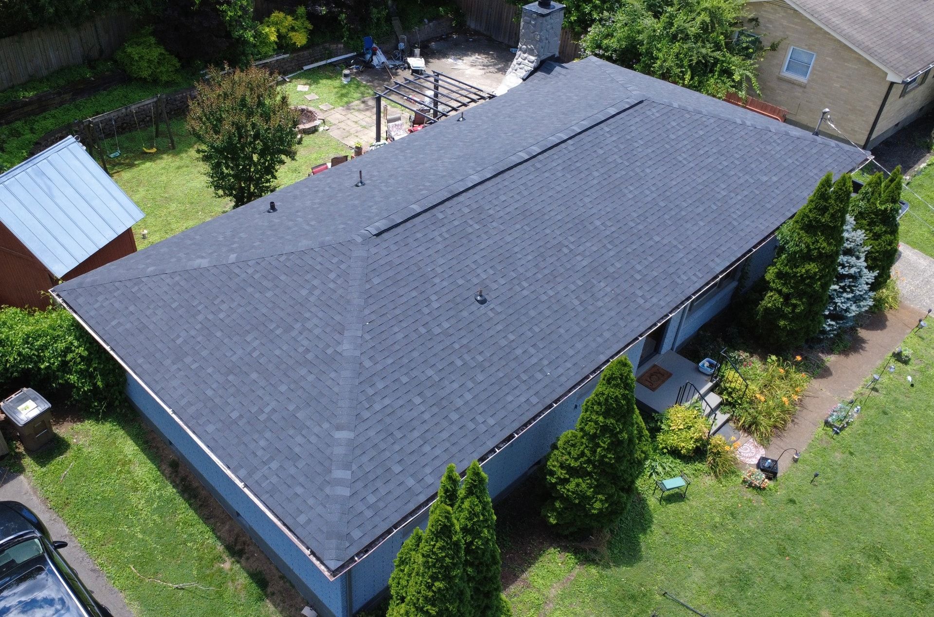 Aerial photo of a single story blue painted brick ranch home with a new charcoal shingle hip roof, a stone chimney, a continuous ridge vent, and tall evergreen shrubs along the front