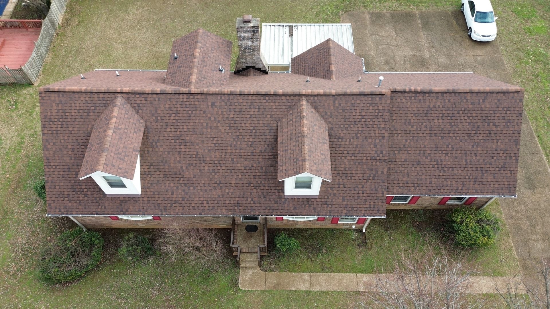 Straight down aerial photo of a brick home with red shutters, two front dormers, a brick chimney, new brown dimensional shingles, and a white metal carport roof