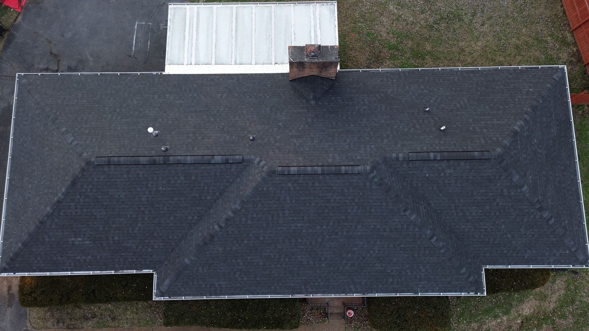 Straight down aerial photo of a long brick ranch home with new charcoal architectural shingles, hip ends, three sections of ridge vent, a brick chimney, and a white metal patio cover at the back