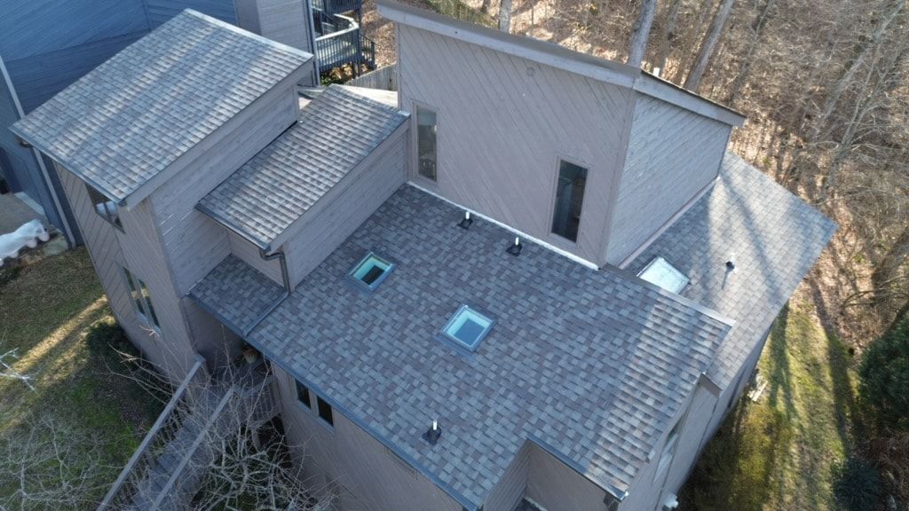 Aerial photo of a contemporary home with diagonal wood siding, a low slope shingle roof with two skylights, stepped upper roof sections, and a wooded hillside behind