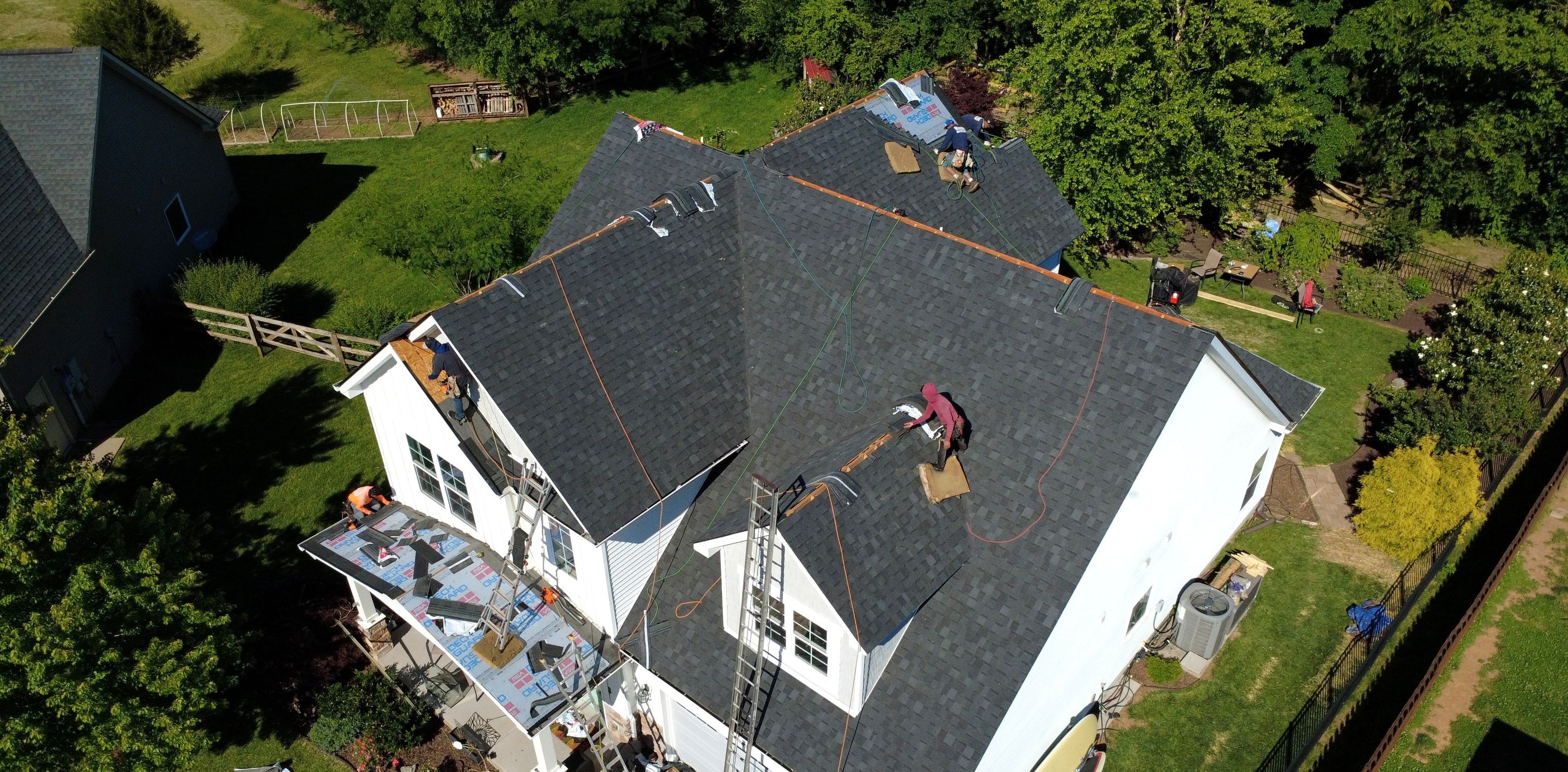 Aerial photo of a crew installing charcoal shingles on a two story white farmhouse, with printed synthetic underlayment visible on the porch roof and ladders against the house