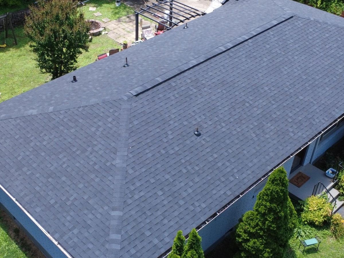 Close aerial view of a hip roof with charcoal architectural shingles, hip caps, pipe boots, and a continuous ridge vent