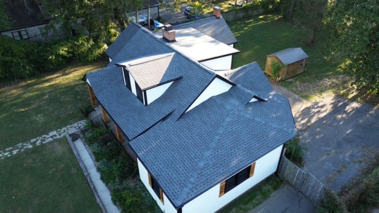 Aerial photo of a white board and batten home with new charcoal architectural shingles, several intersecting gables, a brick chimney, and a covered porch with wood posts
