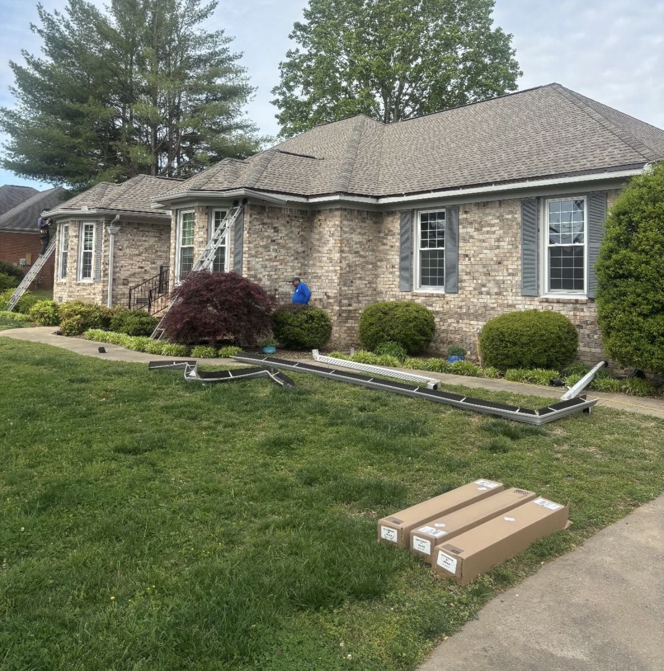 Single story brick home with new seamless gutter sections and downspout boxes staged on the lawn while a crew member works at the eave