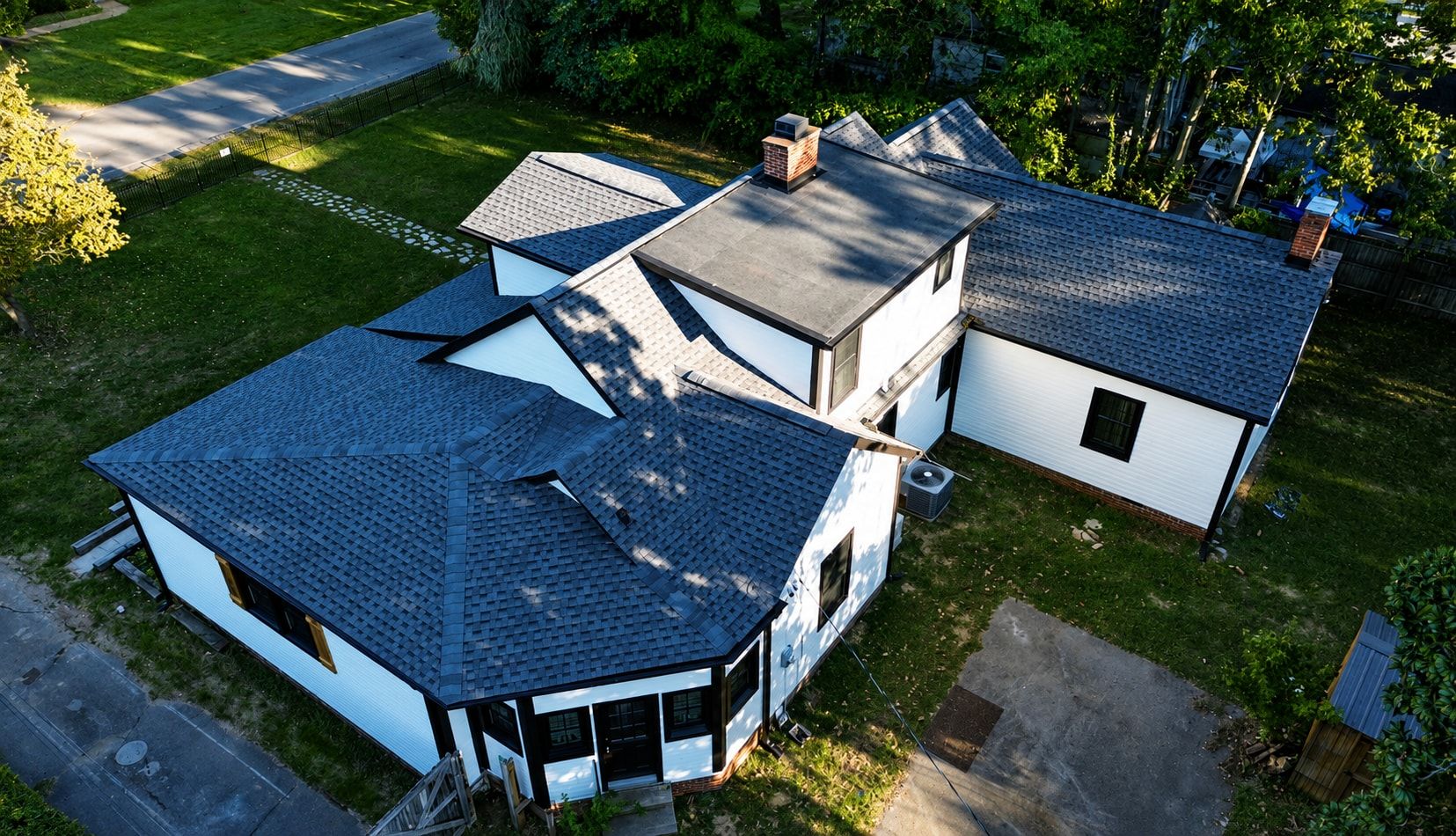 Aerial photo of a white home with new charcoal shingles across several intersecting roof sections, a brick chimney, and a dark low slope membrane roof over a two story addition