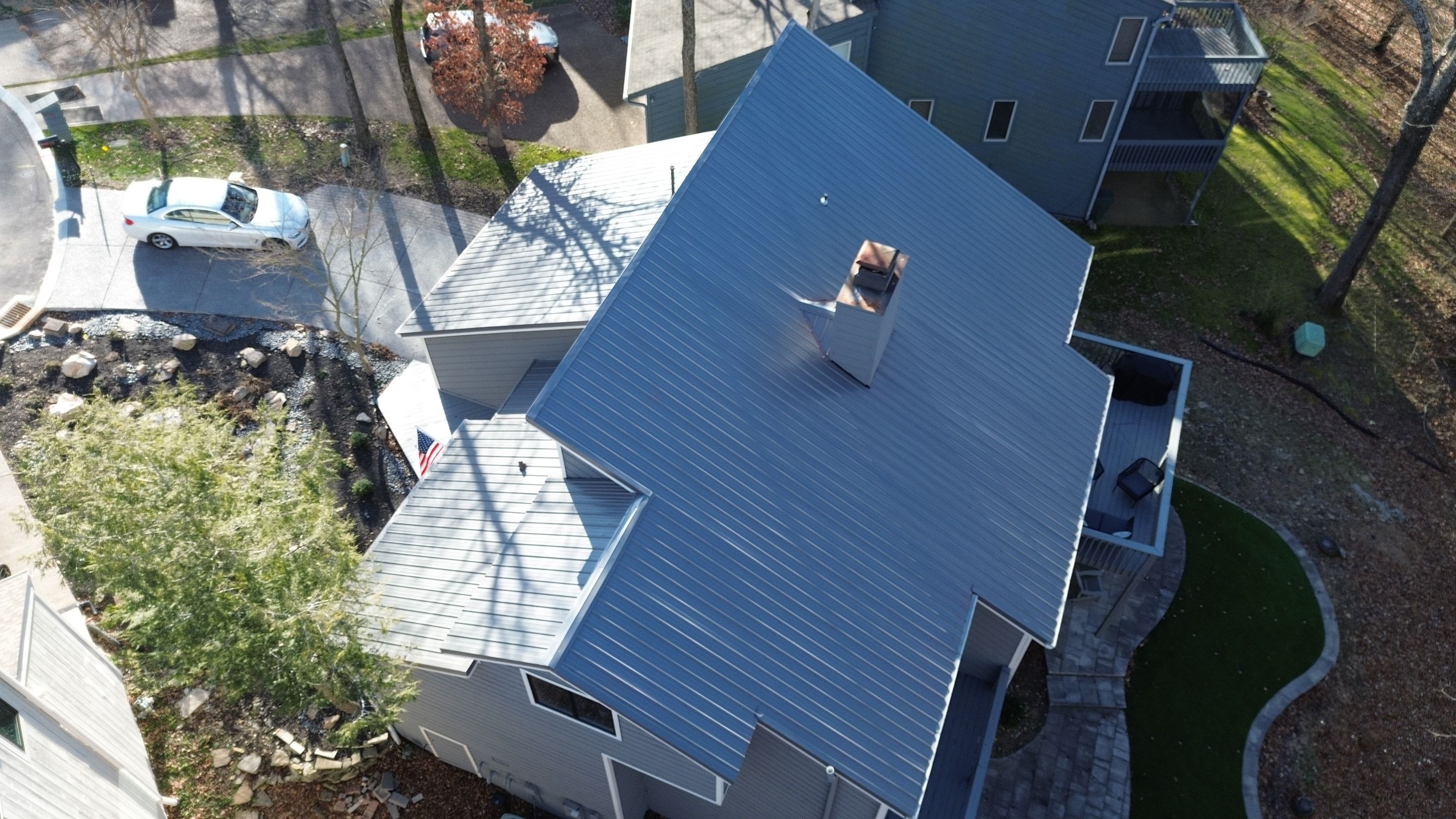 Aerial photo of a charcoal standing seam metal roof on a steep pitched two story home with a chimney, driveway and mature trees around it