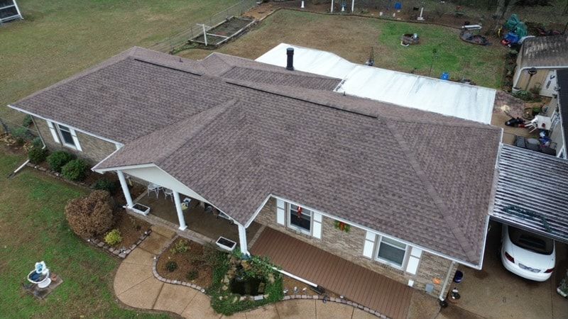 Aerial photo of a single story brick ranch with new weathered wood colored architectural shingles, a gabled front porch with white columns, a carport, and a white metal cover at the back